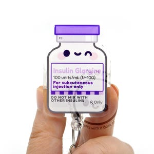 May include: A purple and white acrylic badge reel shaped like an insulin vial. The label reads Insulin Glargine, 100 units/mL (U-100), For subcutaneous injection only, and DO NOT MIX WITH OTHER INSULINS. It features a cute winking face.