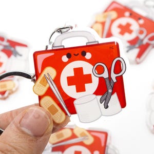 May include: A red first aid kit keychain with a white cross, bandages, scissors, and tape. The keychain has a silver ring and a handle. The design is a cartoon style.