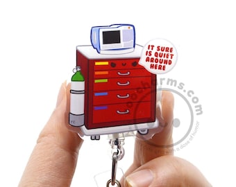 May include: A red medical crash cart badge reel featuring a white oxygen tank on the side and a monitor on top. The cart has a cartoon face and a speech bubble that reads, IT SURE IS QUIET AROUND HERE. The design is printed on a clear acrylic base.
