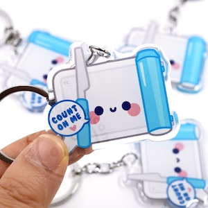 May include: Clear acrylic keychain featuring a cute, smiling design with pink cheeks and a blue speech bubble that says "Count on Me." The keychain has a silver keyring and chain, and a light blue and white color scheme.