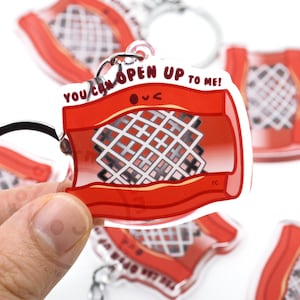 May include: A clear acrylic keychain featuring a red and white design with the text "YOU CAN OPEN UP TO ME!". The keychain has a silver keyring and a cartoon-style illustration.