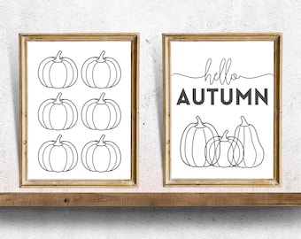 May include: Two framed wall art prints with gold frames. One print features six outlined pumpkins. The other print says "hello AUTUMN" above three outlined pumpkins. The prints are displayed on a wooden shelf.