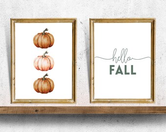 May include: Two framed prints, one with a simple "hello fall" text in a dark green color and the other with three watercolor pumpkins in shades of orange and pink.