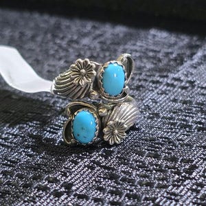 May include: A silver ring featuring two oval-shaped turquoise stones set in serrated bezels. The band is decorated with embossed floral and fan-like patterns on both sides of the stones, creating a symmetrical design with a polished finish.