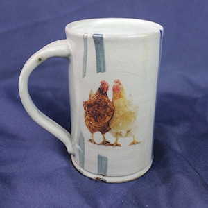 May include: A white ceramic mug with a blue and gray striped design. The mug features a printed image of two chickens, one brown and one white, standing side by side.