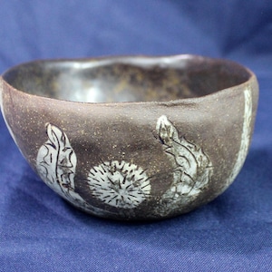May include: A small, brown ceramic bowl with a white design of stylised plants and flowers. The bowl is handmade and has a rustic, earthy feel.