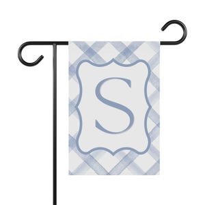Puede incluir: Una bandera de jardín con un patrón de cuadros de acuarela azul claro. La bandera tiene un centro blanco con un borde decorativo azul claro y la letra "S". La bandera se muestra en un soporte de metal negro.