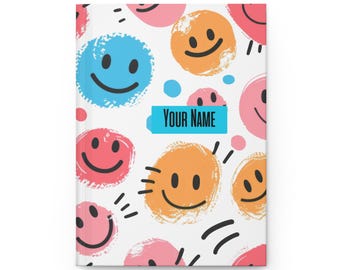 May include: A white notebook cover with a colorful pattern of smiley faces. The smiley faces are in various colors, including blue, orange, pink, and yellow. The text "YOUR NAME" is printed in blue on a white rectangle.