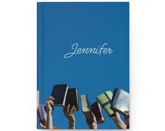 Op de afbeelding: Een blauw notitieboek met de naam "Jennifer" in cursief schrift. Het notitieboek wordt voor een blauwe lucht gehouden, met verschillende handen die boeken vasthouden.