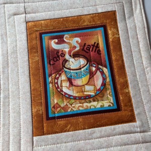 Peut inclure: Un set de table carré matelassé avec une image centrale d'une tasse de café fumante et les mots "Cafe Latte". Le design comprend une bordure marron et bleue, avec un cadre extérieur beige. Le set de table semble fait à la main.