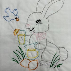 May include: Embroidered design of a white bunny holding a pitcher and glass of lemonade, with a blue bird flying above and lemons on the ground.