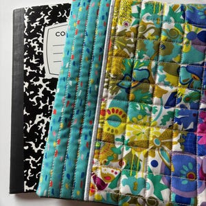 May include: A composition notebook with a black and white cover is next to a quilted fabric case. The case has a teal blue panel with colorful stitching and a floral patchwork design in shades of blue, green, and yellow.