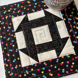 May include: Square quilted coaster with a black background and colorful polka dots. The center features a geometric design in black and white with colorful stitching. A gray mug with the text "Sewing Mends the Soul" rests on the coaster.