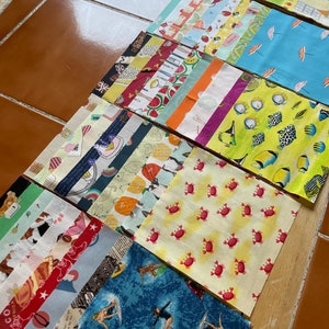May include: A collection of fabric squares with various patterns, including crabs, surfers, and food items. The fabrics are arranged on a brown surface, showcasing a variety of colors and designs suitable for quilting or crafting projects.