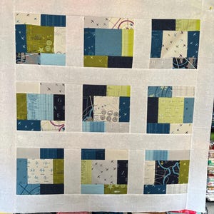 May include: A quilt top featuring a grid of nine square blocks separated by white sashing. Each block is composed of smaller rectangular fabric pieces in shades of blue, green, and gray, featuring patterns of airplanes, maps, and grids.