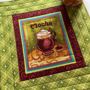 Peut inclure: Un set de table vert matelassé avec une image centrale d'un moka avec de la crème fouettée et le mot "Mocha". Le set de table a un motif à carreaux et une bordure décorative. Une tasse en céramique avec un motif floral est dans le coin supérieur droit.