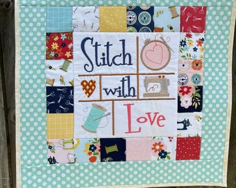 May include: A light blue and white polka dot fabric square with a white border. The center of the square features a patchwork design with various fabrics and a white embroidered design that says "Stitch with Love".