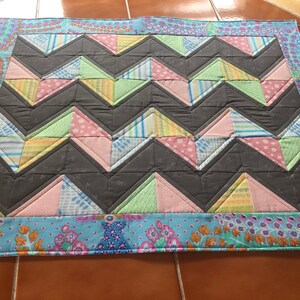 May include: A gray, pink, green, and blue patchwork quilt with a chevron pattern. The quilt has a floral border.