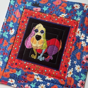 May include: A square, quilted fabric item featuring a colorful cartoon dog in the center. The dog has a black nose, big eyes, and a multi-colored body. The dog is set against a black square, surrounded by red and blue floral patterned fabric.