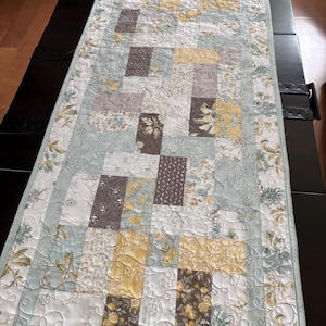 May include: A rectangular quilted table runner featuring a patchwork design of rectangular fabric blocks in shades of sage green, soft yellow, white, and dark brown, accented with intricate floral patterns and swirling machine quilting.