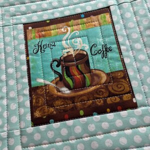 May include: A quilted coffee mug mat with a central square featuring a coffee cup design. The cup is brown with colorful stripes, steam rising, and the words "Hona Coffee". The mat has a teal background with white polka dots.