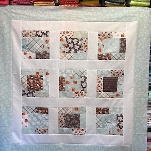 May include: A handmade quilt with a patchwork design. The quilt features squares with autumn-themed patterns, including pumpkins and leaves, in shades of blue, brown, and white. The quilt is framed by a light blue floral border and a white inner border.