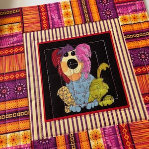 May include: A colorful, quilted placemat featuring a cartoon dog in pajamas. The dog is set against a black background with a red border. The outer border has a patchwork design in shades of orange, purple, and yellow.