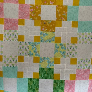 May include: A quilt with a patchwork design featuring squares of yellow, pink, green, and white fabric. The squares are arranged in a grid pattern, creating a geometric design.