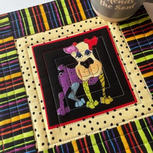May include: A colorful quilted placemat featuring a cartoon dog with a heart-shaped nose and a polka-dotted background. The placemat has a black border with vertical stripes in various colors. A gray mug with the text "Sewing Mends the Soul" is on the placemat.