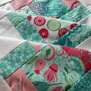 May include: Close-up of a colorful quilt block featuring sea creatures and patterns. The design incorporates pink, teal, and white fabrics with images of jellyfish, fish, and other marine life. The quilt block is made of various fabric pieces.