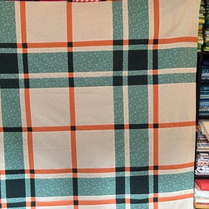 Peut inclure: Un tissu crème à motif écossais, avec des lignes orange et noires, et des sections imprimées florales turquoise. Ce tissu est probablement destiné au quilting ou à la couture. L'arrière-plan montre des étagères de tissus pliés.