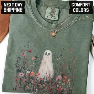 May include: A moss green t-shirt featuring a ghost illustration in a field of colorful flowers. The shirt has a crew neck and short sleeves. A gold necklace with a small pendant is draped on the shirt.