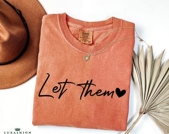 May include: A coral colored t-shirt with the text "Let Them" in black script with a small black heart to the right of the text.