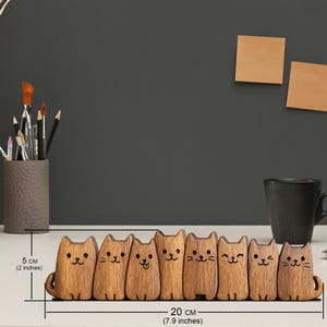 Puede incluir: Un organizador de escritorio de madera con forma de gato, de 20 cm de largo y 5 cm de alto, con siete figuras de gato individuales. Cada gato tiene una expresi&oacute;n facial &uacute;nica. Tambi&eacute;n se ven un portal&aacute;pices y una taza.