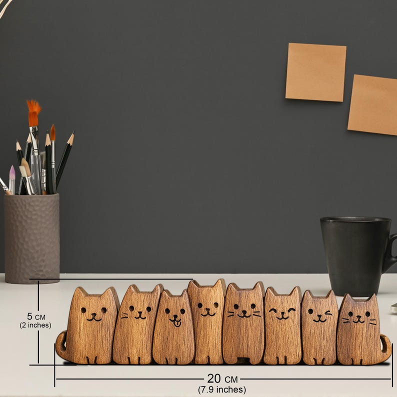Puede incluir: Un organizador de escritorio de madera con forma de gato, de 20 cm de largo y 5 cm de alto, con siete figuras de gato individuales. Cada gato tiene una expresi&oacute;n facial &uacute;nica. Tambi&eacute;n se ven un portal&aacute;pices y una taza.