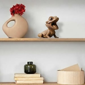 May include: A decorative shelf arrangement featuring a beige vase with a red floral arrangement, a wooden lizard figurine with an open mouth, a green glass bottle, stacked books, and a wooden box. The items are arranged on wooden shelves against a white wall.