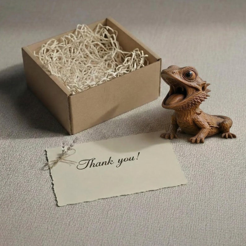 May include: A detailed wooden figurine of a bearded dragon with its mouth open, next to a small cardboard box filled with packing material. A card with the words "Thank you!" is also present.