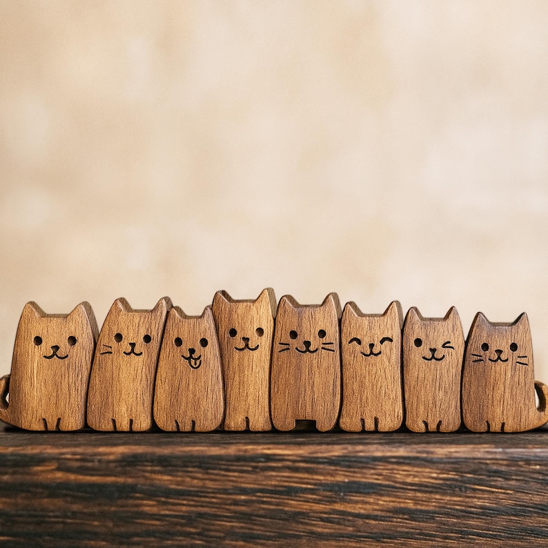 Puede incluir: Una fila de ocho figuras de gato de madera con diferentes expresiones faciales. Cada gato est&aacute; tallado en madera de color marr&oacute;n claro y tiene detalles negros simples para los ojos, la nariz y los bigotes. Los gatos est&aacute;n colocados sobre una superficie de madera oscura.