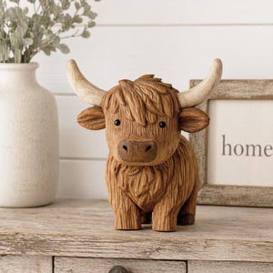 May include: A brown Highland cow figurine with textured fur, large horns, and small black eyes. The figurine is standing on a wooden surface, with a vase and a framed sign that says "home" in the background.