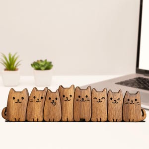 May include: A row of eight wooden cat figurines with engraved facial features and varying expressions. The cats are light brown and stand upright, with a small tail on each. The figurines are arranged on a white surface.