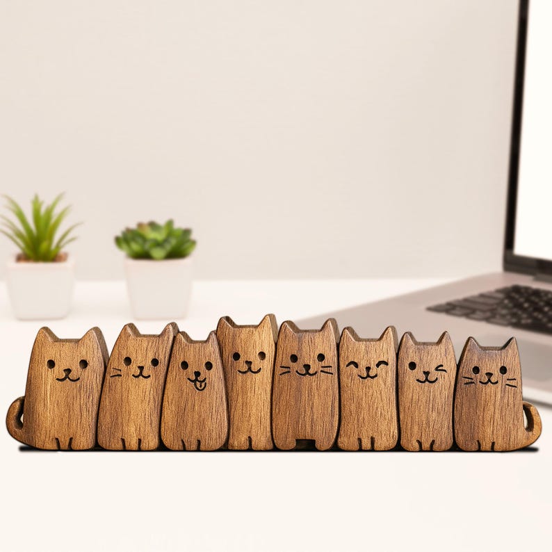 Puede incluir: Ocho figuras de gatos de madera con caras grabadas y diferentes expresiones. Los gatos de color marr&oacute;n claro est&aacute;n de pie en fila. Cada uno tiene una peque&ntilde;a cola. Se exhiben sobre una superficie blanca, con un port&aacute;til y peque&ntilde;as plantas.