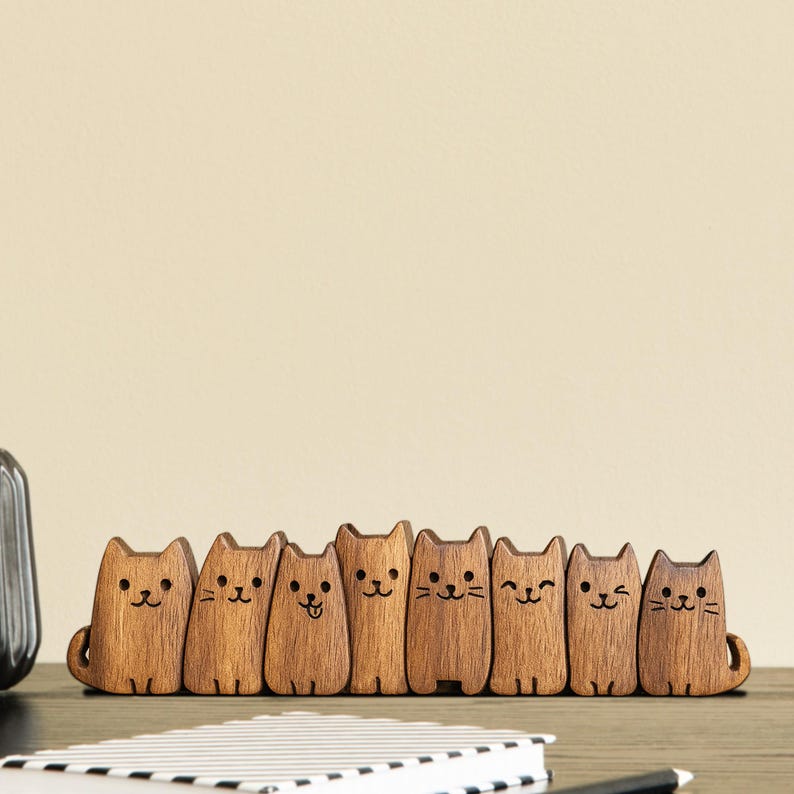 Puede incluir: Una fila de ocho figuras de gatos de madera con rasgos faciales grabados y peque&ntilde;as colas. Los gatos son de varios tonos marrones y est&aacute;n de pie, creando una exhibici&oacute;n decorativa caprichosa.