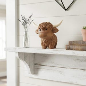 May include: A brown Highland cow figurine with cream-colored horns. The decorative sculpture has a textured, shaggy coat and is displayed on a white shelf. The shelf also holds a glass vase with lavender and a stack of books.