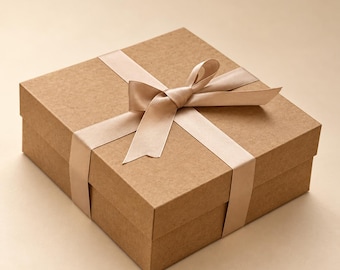 May include: A square gift box made of light brown cardboard with a smooth matte finish. The box is wrapped with a wide, light beige satin ribbon tied into a centered bow on the top lid, featuring clean edges and a simple, structured design.