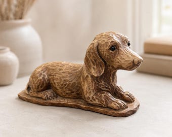 May include: A brown dachshund figurine crafted with a carved wood texture, featuring a long body, floppy ears, and dark eyes. The dog is depicted in a resting, lying-down position on an integrated oval base, showcasing detailed, etched fur patterns.