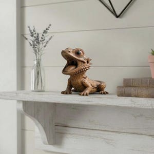 May include: A detailed, brown resin bearded dragon figurine with an open mouth, showcasing its tongue. The figurine has textured skin and is positioned on a white shelf. Decorative home decor.