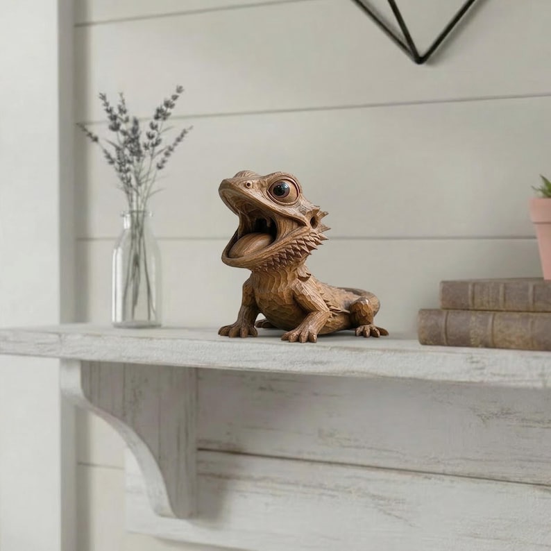 May include: A detailed, brown resin bearded dragon figurine with an open mouth, showcasing its tongue. The figurine has textured skin and is positioned on a white shelf. Decorative home decor.