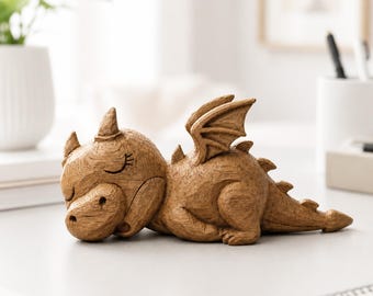 May include: A small, light brown dragon figurine with a textured, wood-like finish. The dragon is depicted in a resting, prone position with its eyes closed, featuring small horns, folded wings, and a ridged tail extending along the surface.