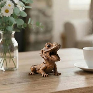 May include: A detailed wooden sculpture of a bearded dragon with its mouth open, revealing its tongue. The lizard is brown with textured details, and its eyes are wide. The sculpture is on a wooden table, with a vase of flowers in the background.