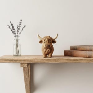 May include: A wooden Highland cow figurine with curved horns and shaggy fur stands on a wooden shelf. A clear glass vase with lavender sprigs and two vintage books are also on the shelf. The background is a plain white wall.